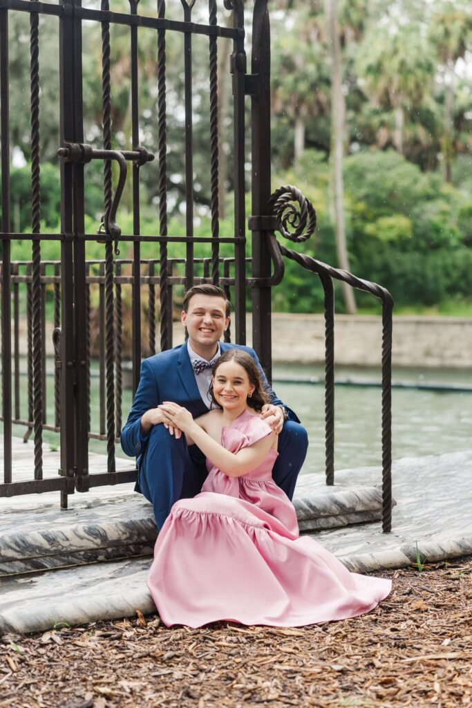 Couple in a pink gown and navy suit smiling on the steps by the iron gates, with the moat behind them
